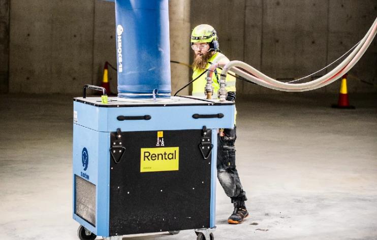 En byggarbetare i varselkläder och hjälm bakom en stor blå luftrenare på hjul på en byggarbetsplats.